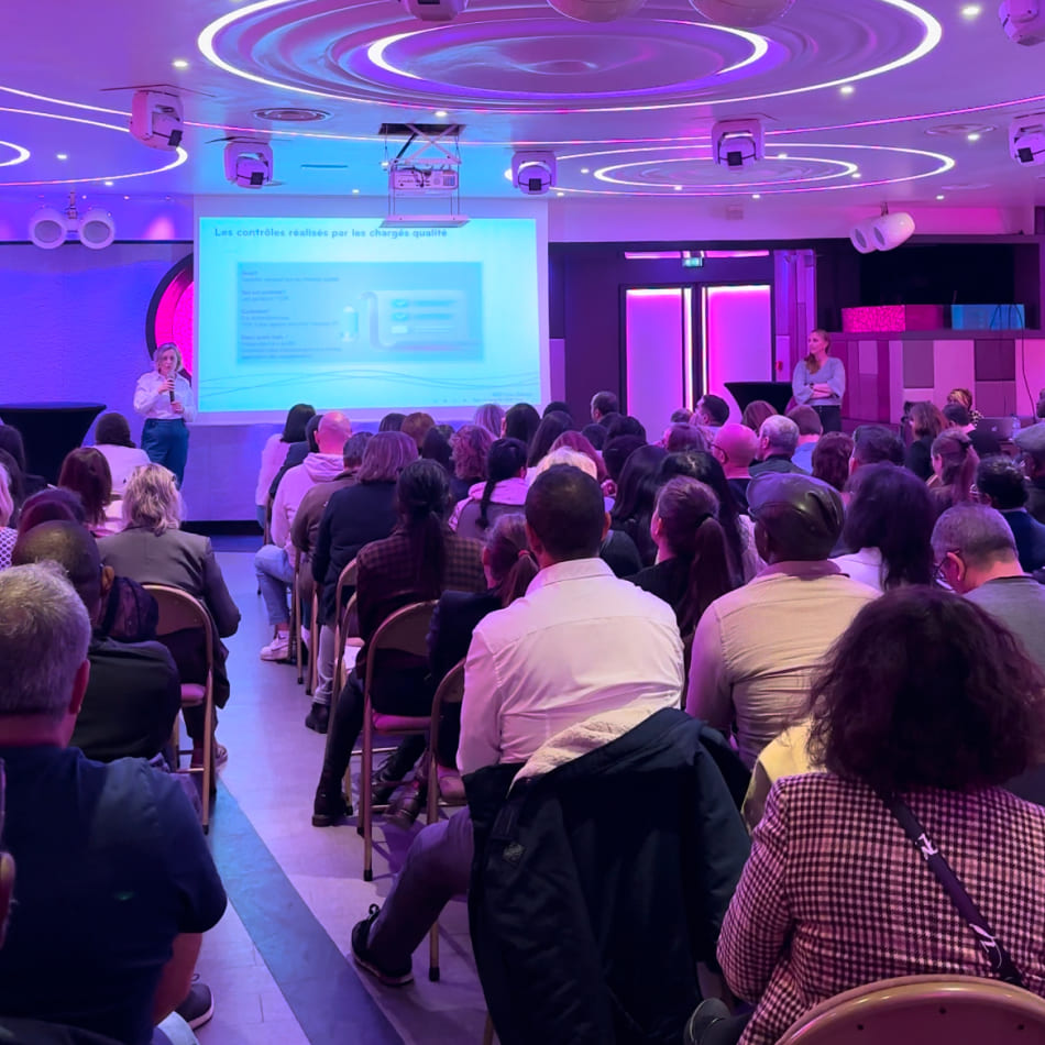 Conférence dans une salle moderne avec une présentatrice devant un écran de projection et une audience attentive.