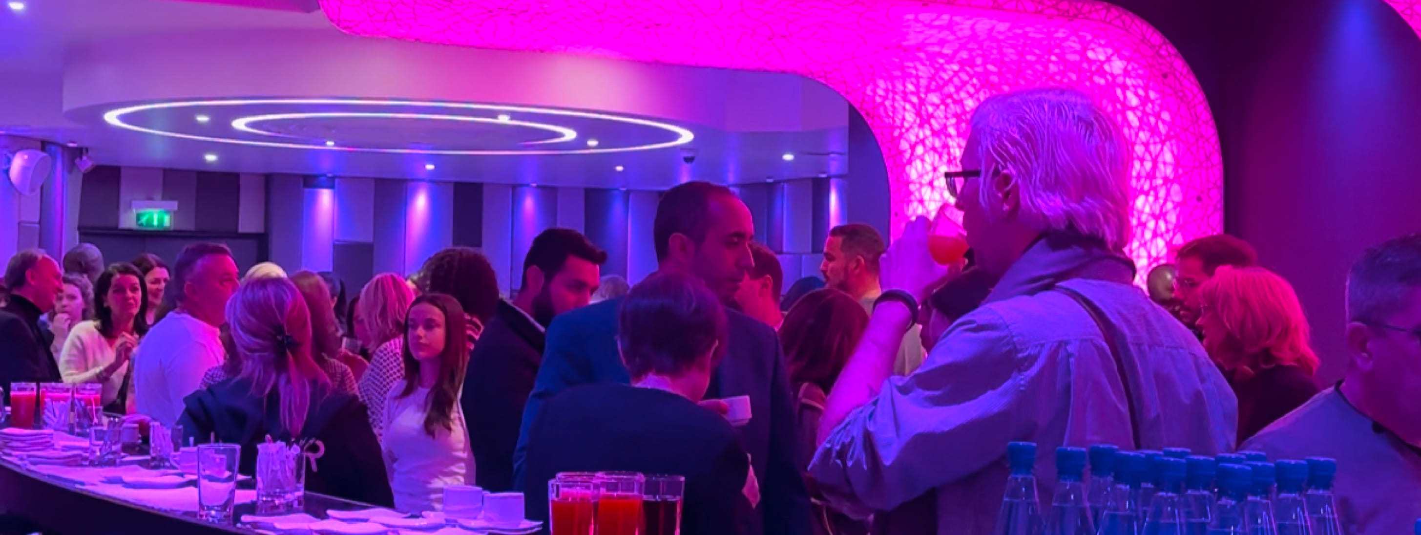 Groupe de personnes discutant et prenant un verre autour d'un bar dans une salle à l'éclairage d'ambiance rose et violet.
