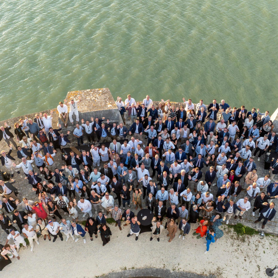 Vue aérienne de nombreux participants rassemblés sur les remparts d'un fort en bord de mer pour une photo de groupe.