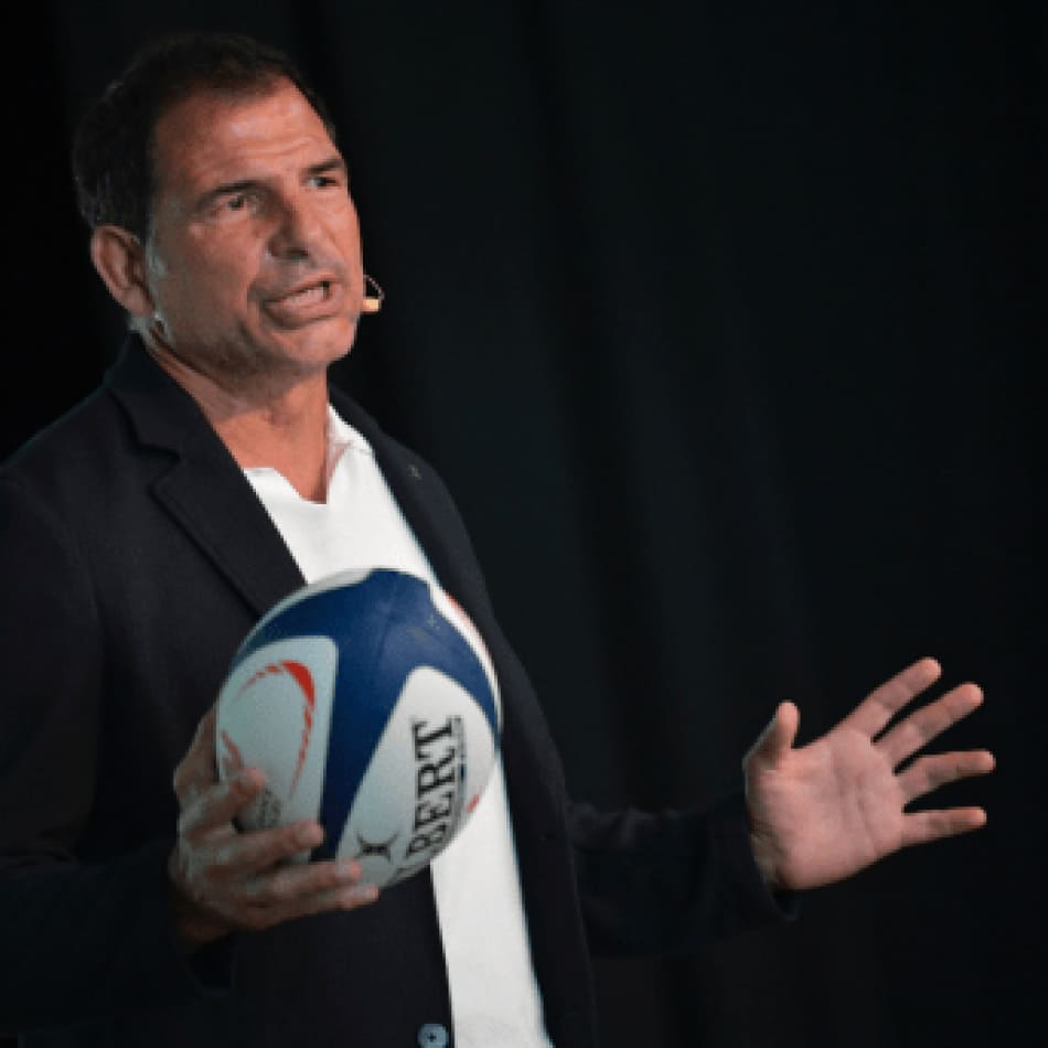 Un intervenant s'exprimant sur scène tout en tenant un ballon de rugby lors d'une conférence.