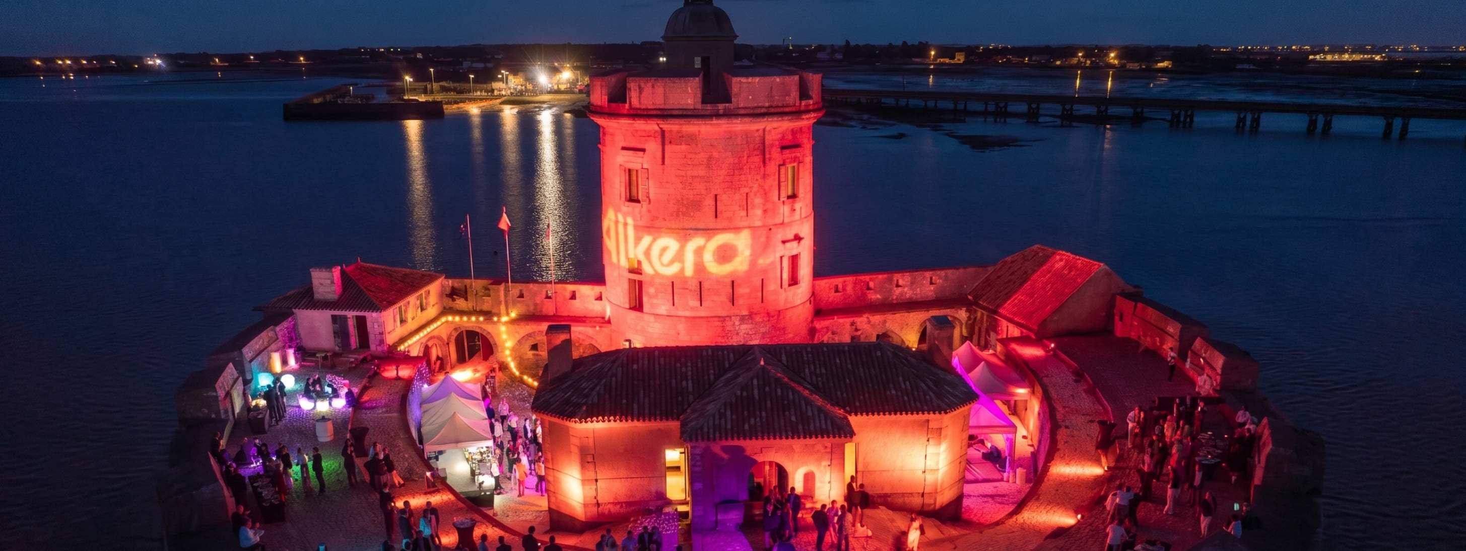 Vue nocturne spectaculaire d'un fort historique illuminé en rouge pour un événement privé avec le logo "Alkera" projeté sur la tour principale.