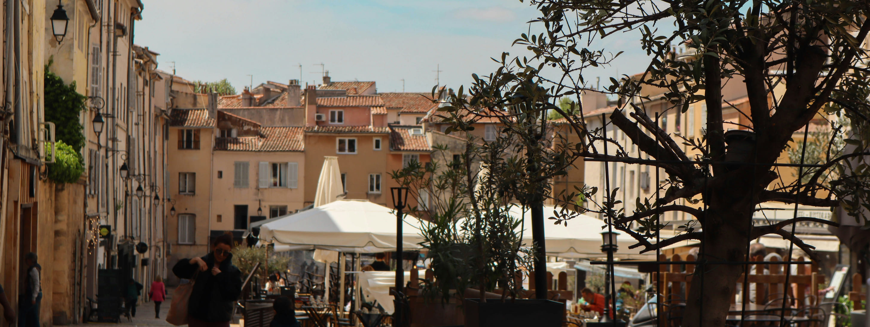 Rue ensoleillée du centre historique d'Aix-en-Provence avec des maisons aux façades ocre, des terrasses de café ombragées par des parasols blancs et un olivier au premier plan.