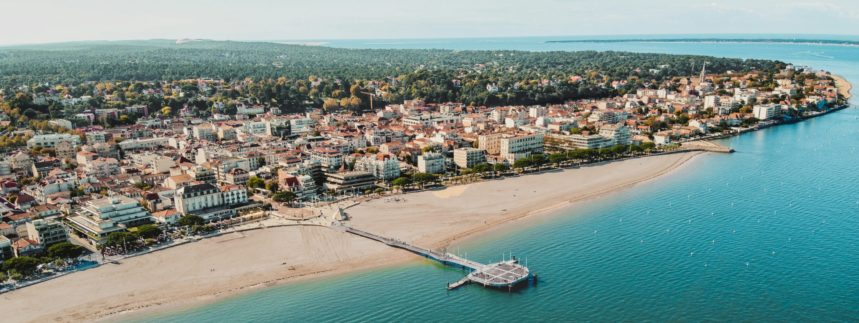 Vue aérienne du front de mer d'Arcachon montrant la plage, la jetée Thiers et les bâtiments de la ville bordés par une forêt.