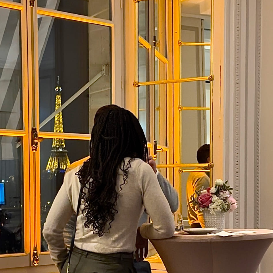 Vue sur la Tour Eiffel illuminée à travers une grande fenêtre, avec une invitée au premier plan lors d'une soirée.