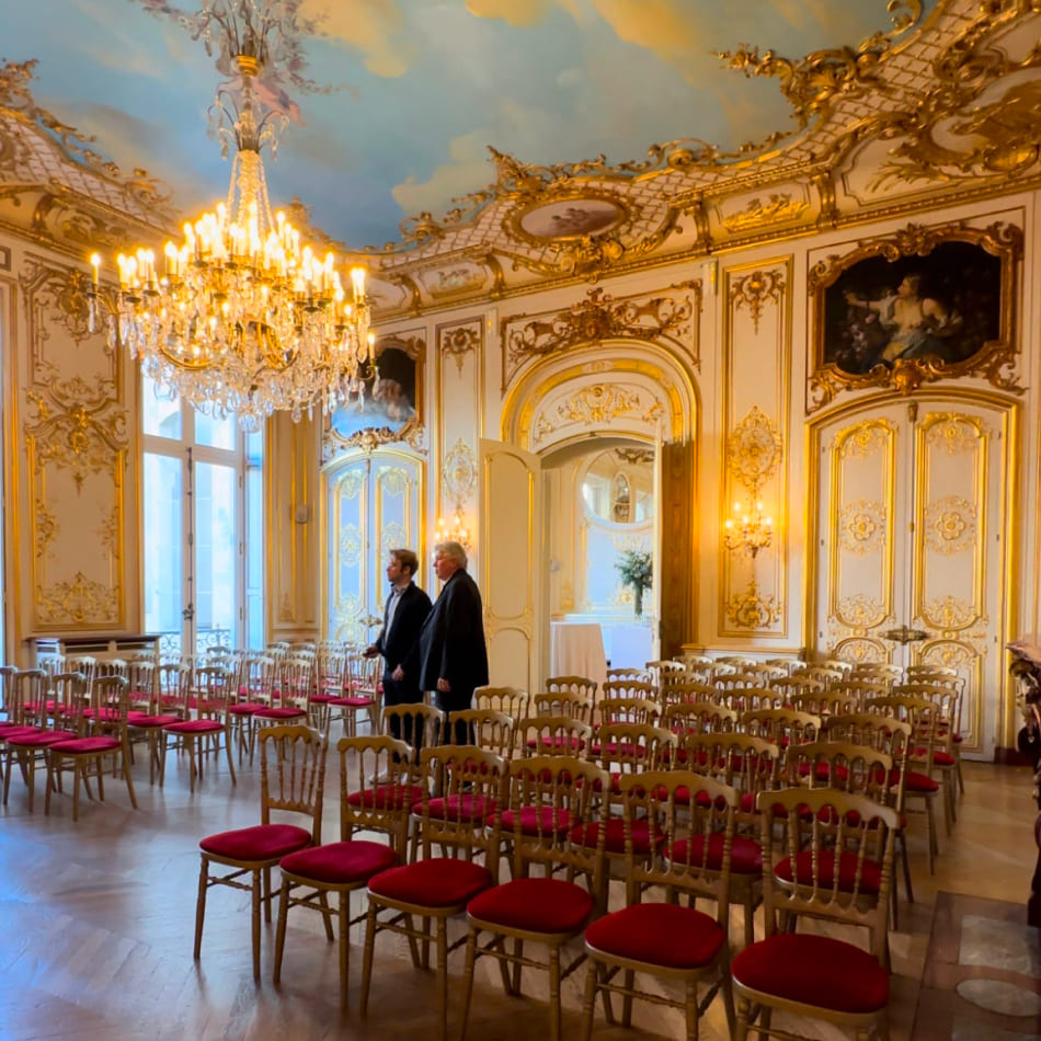 Salle de réception majestueuse avec un grand lustre, des moulures dorées et des rangées de chaises rouges préparées pour un événement.