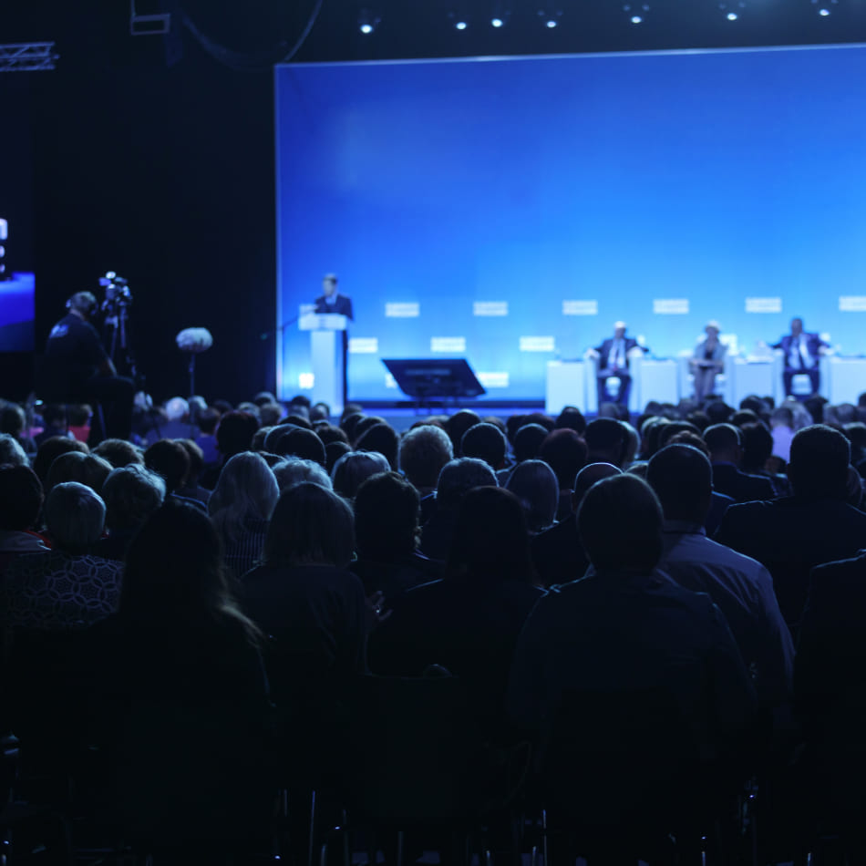 Vue de dos d'une large audience assise dans une salle sombre, orientée vers une scène éclairée d'un bleu vif où se tiennent un orateur et un panel d'intervenants.