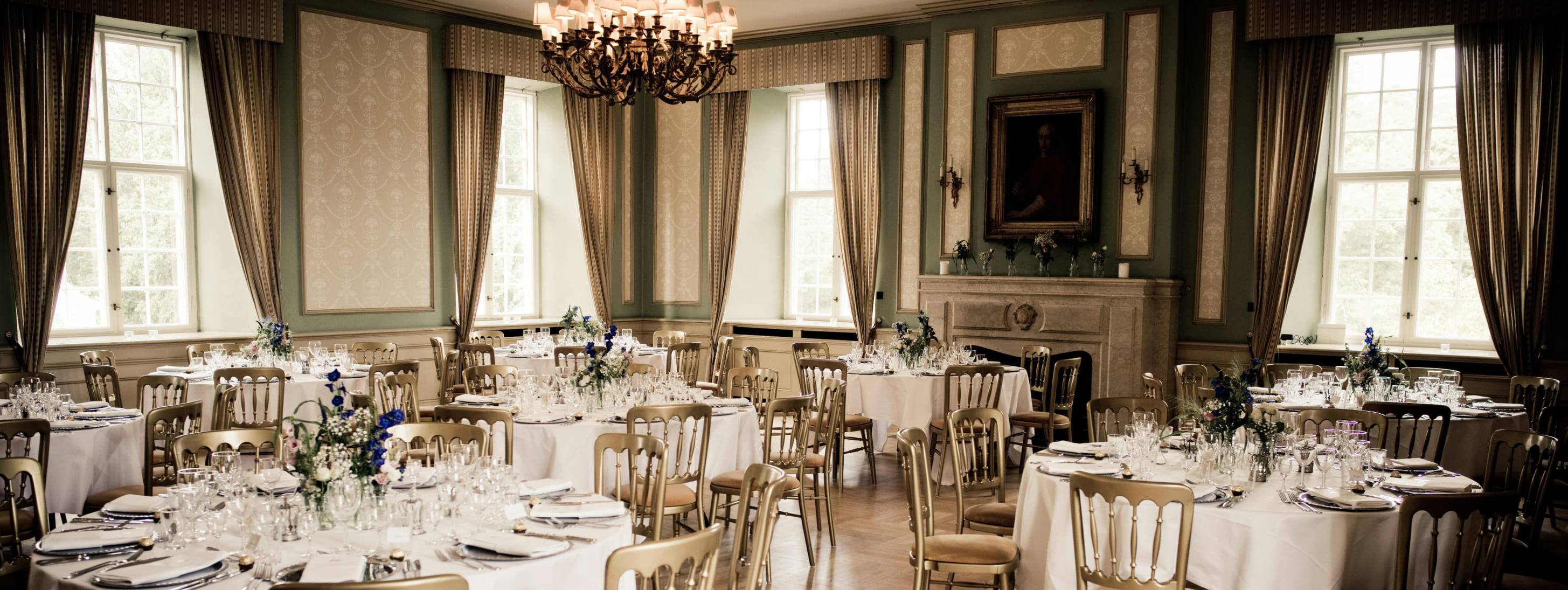 Une élégante salle de réception lumineuse, décorée d'un grand lustre classique, avec des tables rondes dressées de nappes blanches et de chaises dorées.