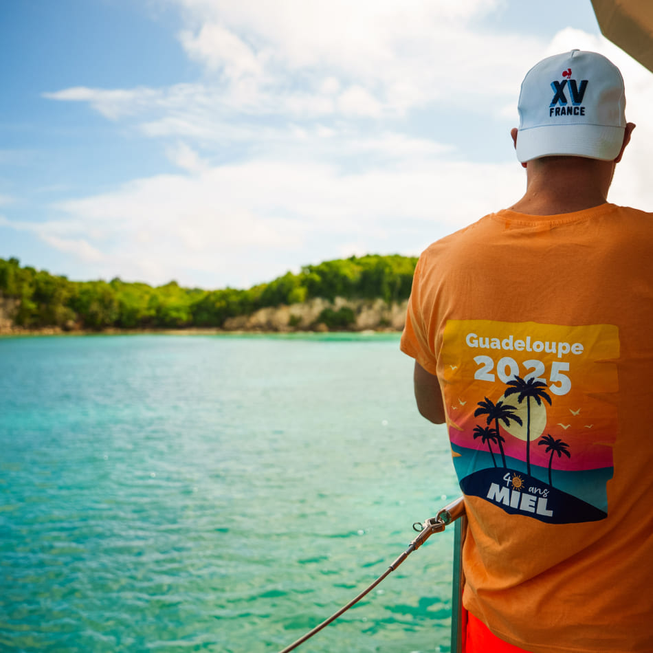 Un homme de dos sur un bateau admire une baie en Guadeloupe, portant un t-shirt orange marqué « 40 ans MIEL ».