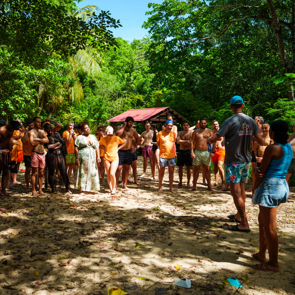 Un groupe de participants en tenue de plage écoute un animateur lors d'un séminaire en plein air sous les arbres.