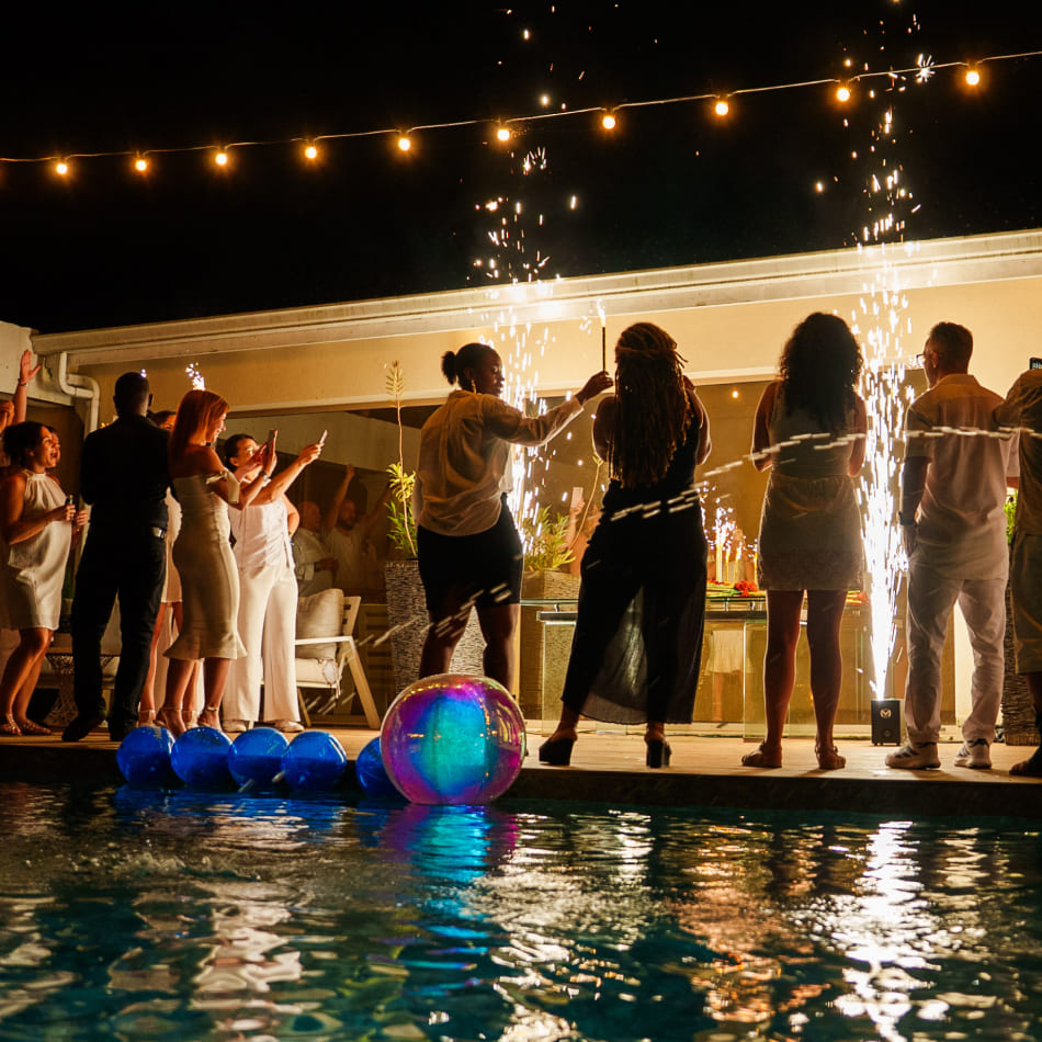 Des invités célèbrent une soirée au bord d'une piscine avec des jets d'étincelles sous une guirlande lumineuse.