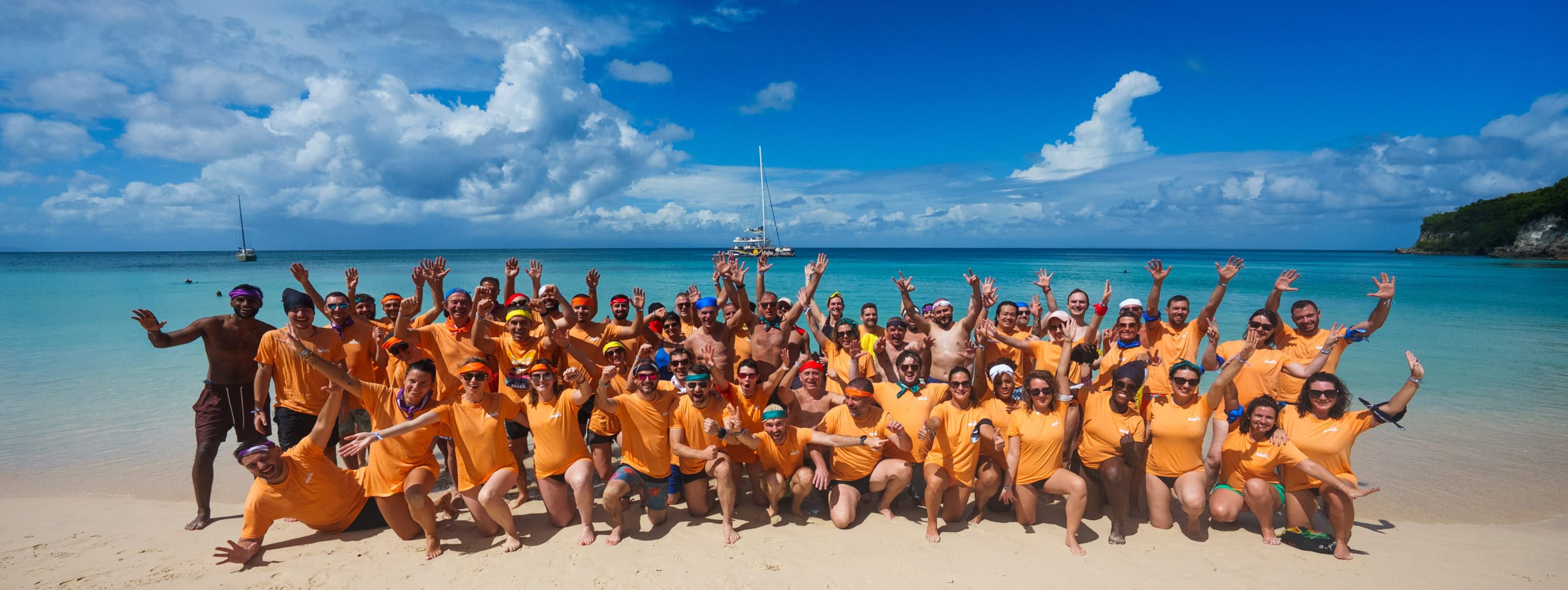 Grande photo de groupe de nombreux participants vêtus de t-shirts orange posant avec enthousiasme sur une plage paradisiaque.