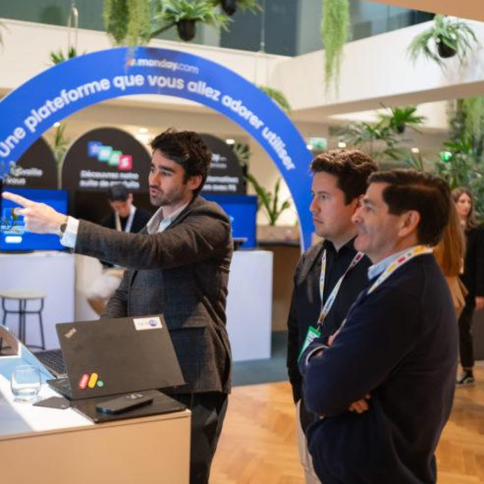 Un présentateur explique les fonctionnalités d'une plateforme de gestion à deux visiteurs sur un stand d'exposition.