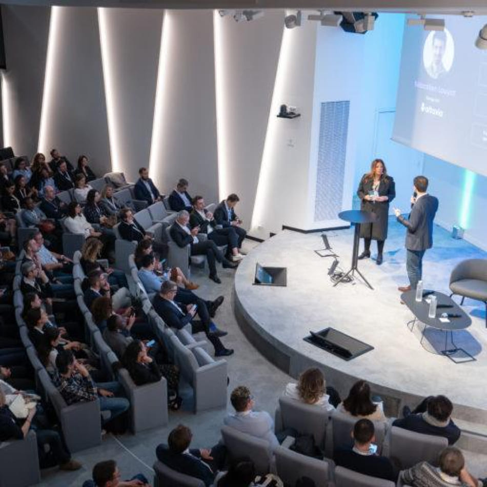 Vue plongeante d'une salle de conférence moderne avec deux intervenants sur scène devant une audience assise.