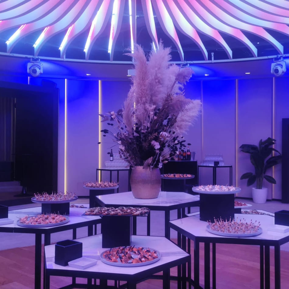 Buffet cocktail haut de gamme dans une salle à l'éclairage bleuté futuriste, avec des tables hexagonales en marbre et une grande composition de pampas au centre sous un plafond design.