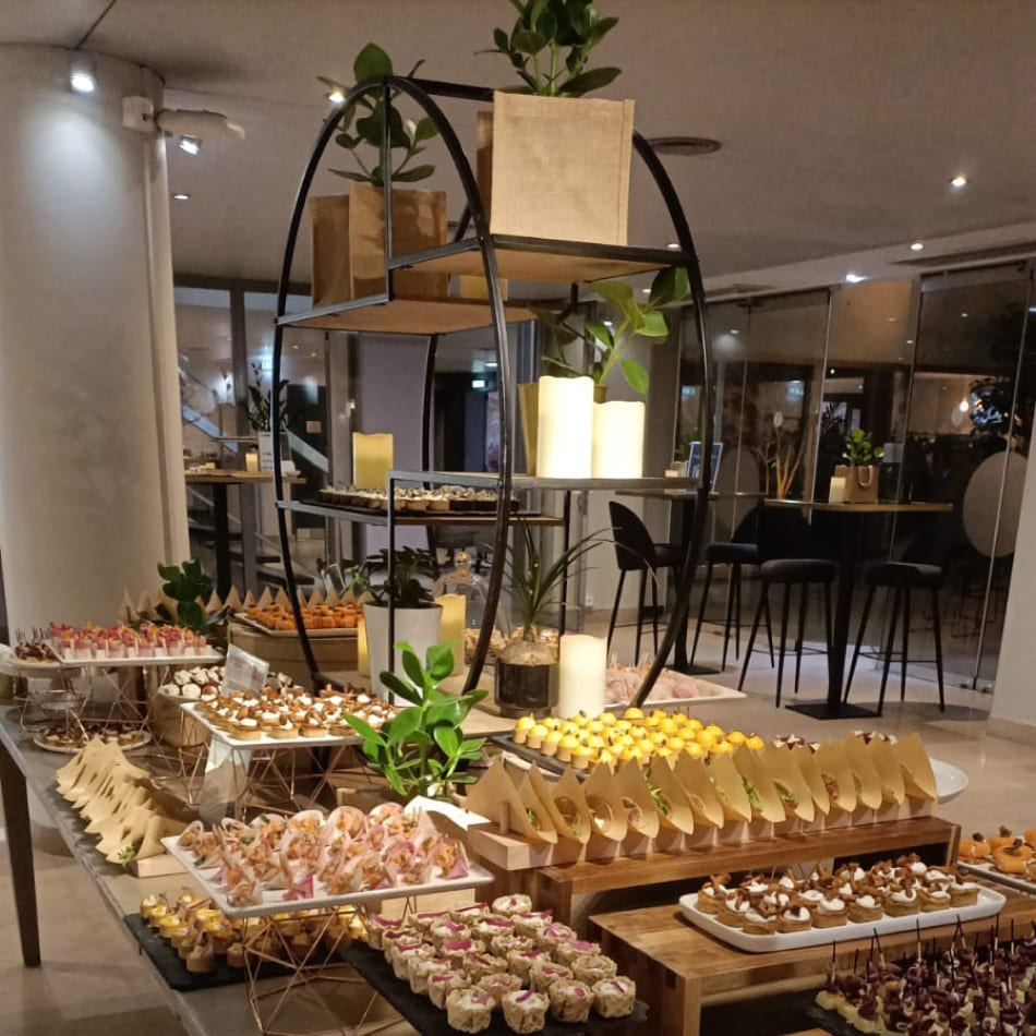 Présentation d'un buffet traiteur varié sur des supports en bois et métal, incluant une étagère circulaire moderne décorée de plantes et de bougies au milieu de mignardises salées et sucrées.