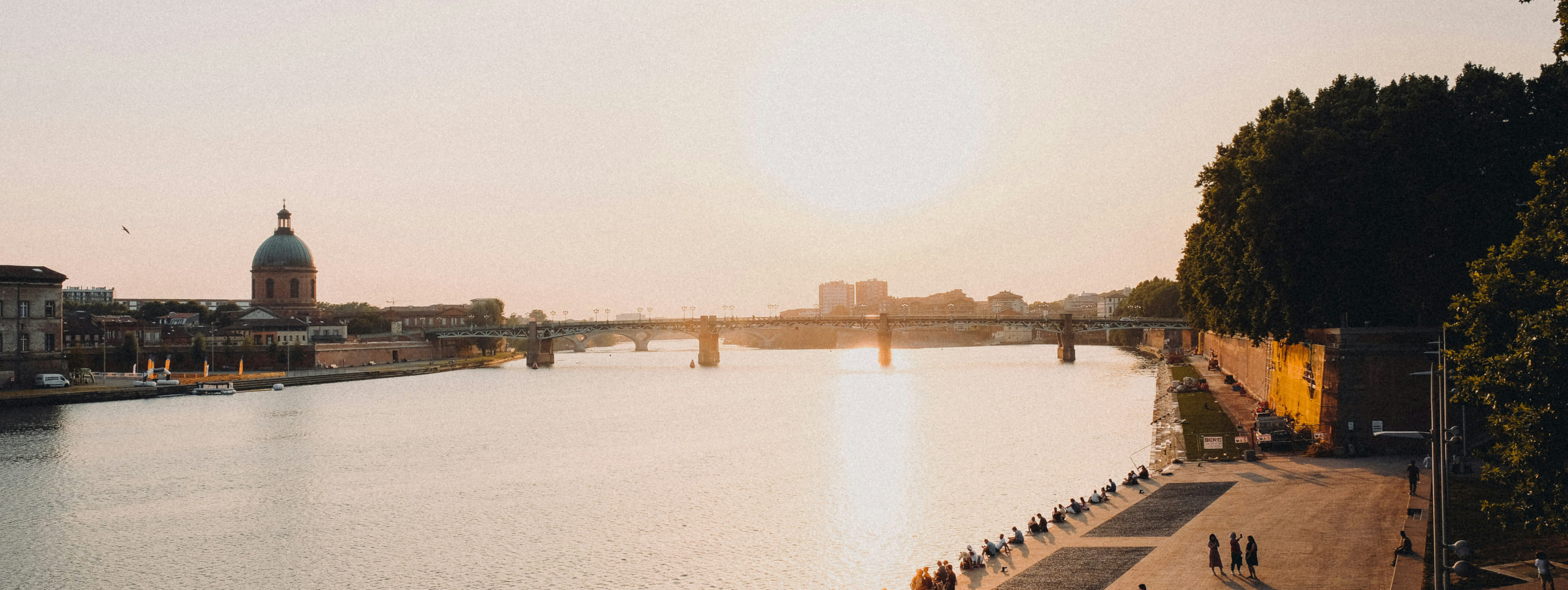 Vue panoramique de Toulouse au soleil couchant, montrant le fleuve Garonne, le dôme de l'Hôpital de la Grave et le Pont Saint-Pierre avec des passants sur les quais.