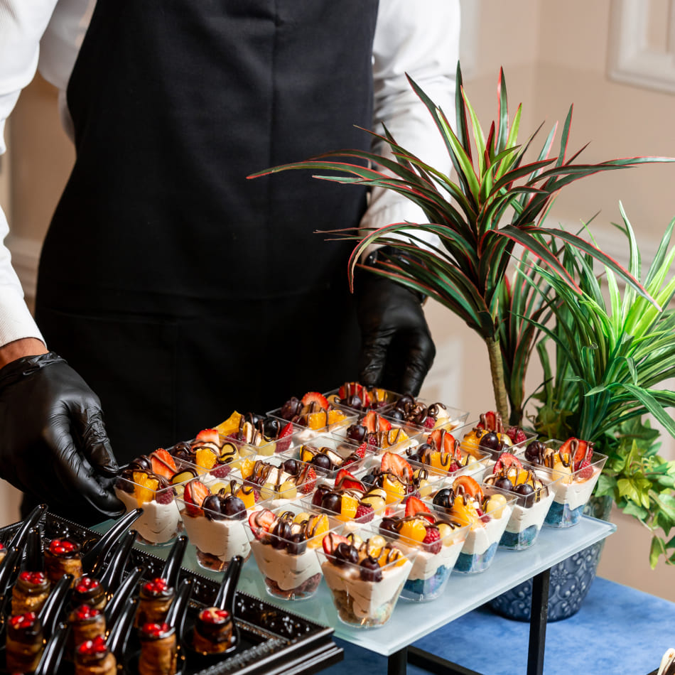 Un serveur en tablier noir et gants de protection présente un plateau garni de nombreuses verrines de desserts aux fruits frais (fraises, raisins, mangues) nappés de chocolat.