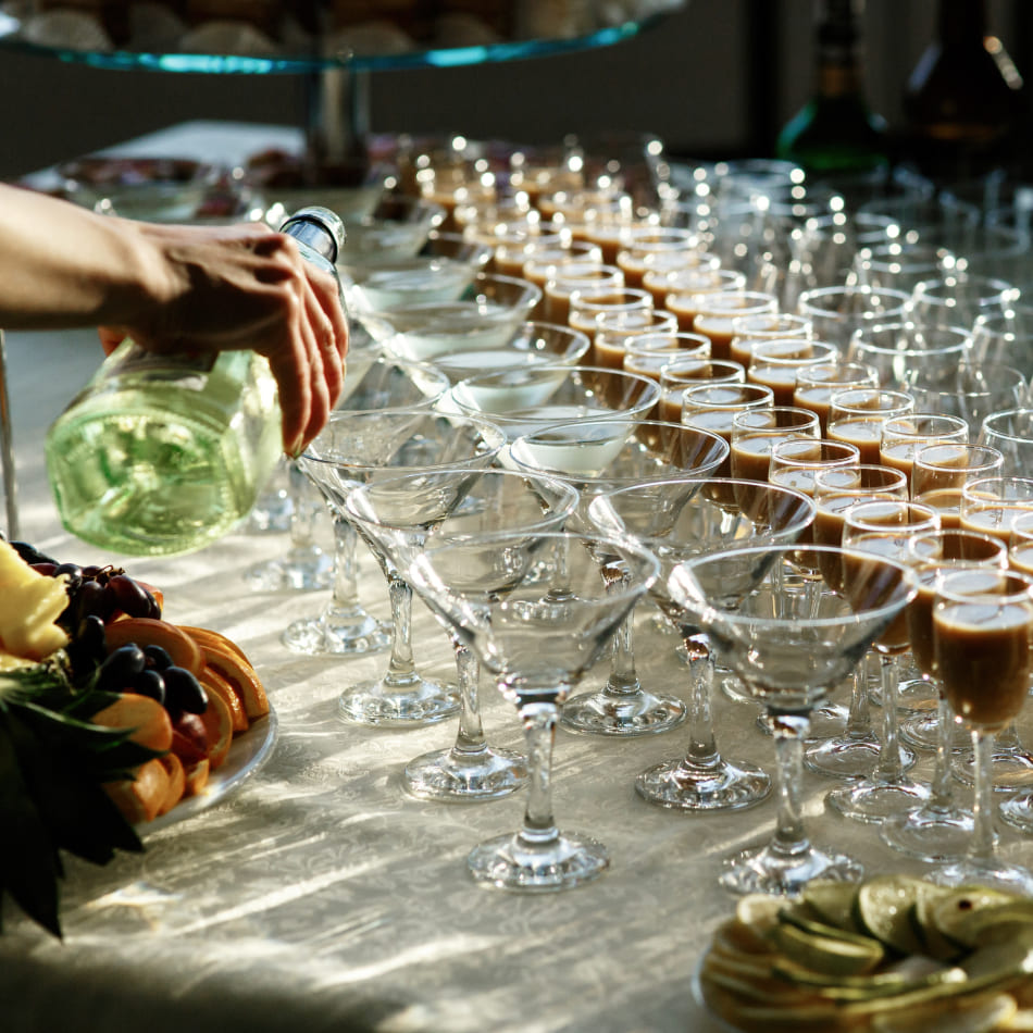 Gros plan sur une main versant une boisson dans des verres à cocktail alignés sur une nappe blanche lors d'un événement.